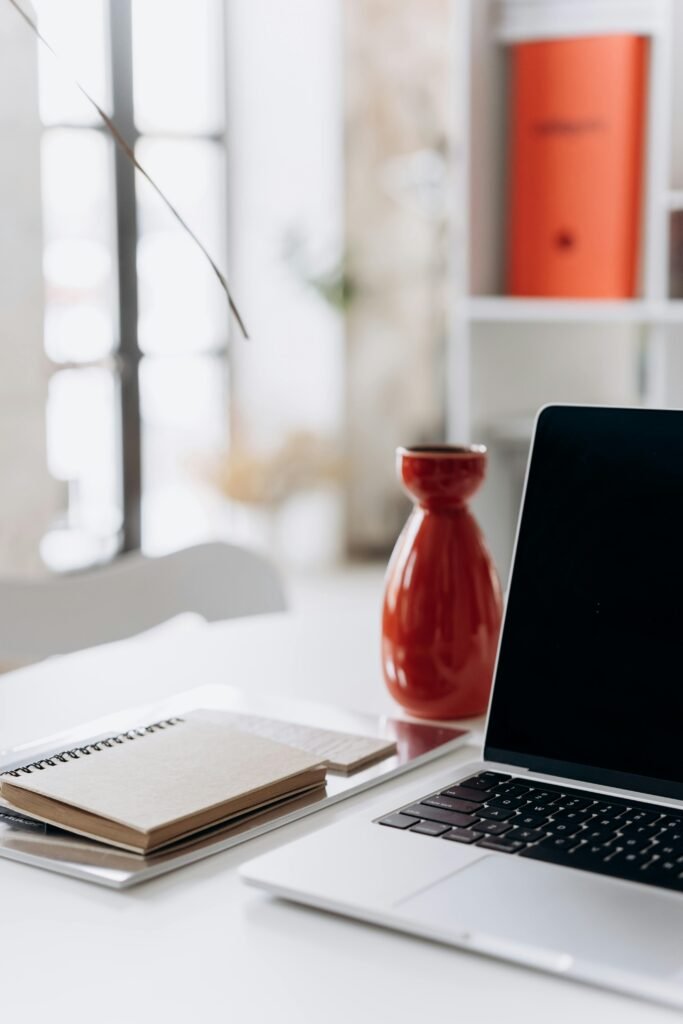 A minimalist home office setup featuring a laptop, notebook, and a red vase on a white desk.